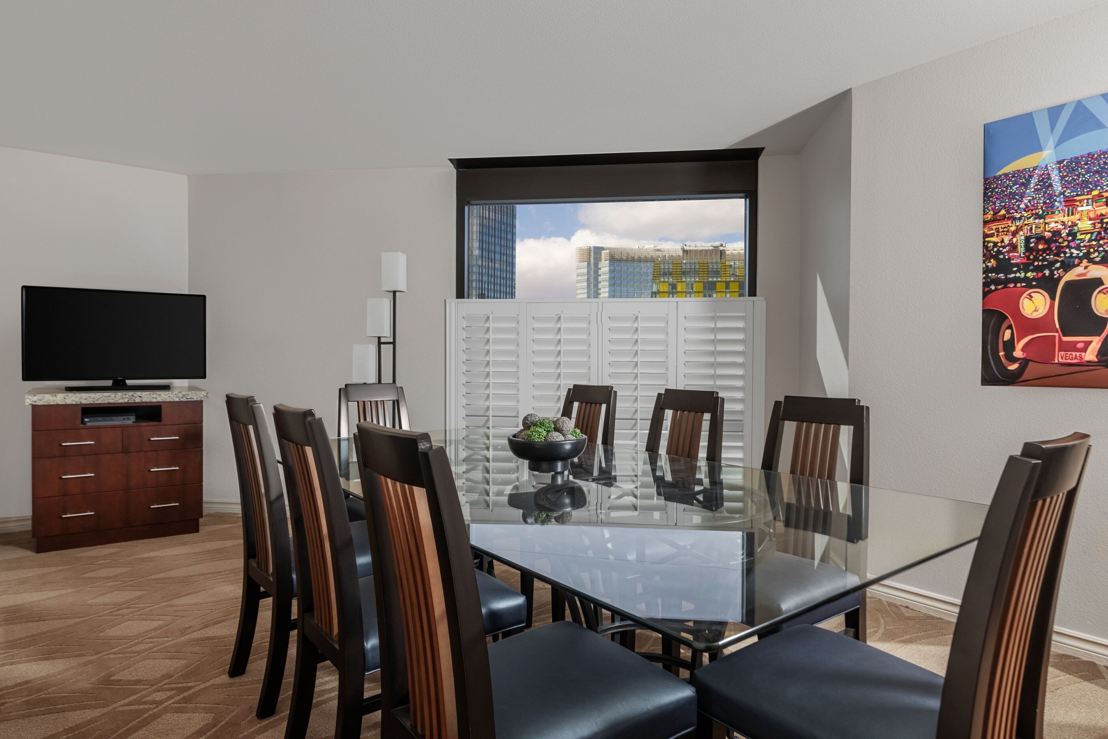 Un comedor con una mesa de vidrio, seis sillas, un televisor sobre un mueble de madera y una ventana con vistas a la ciudad.