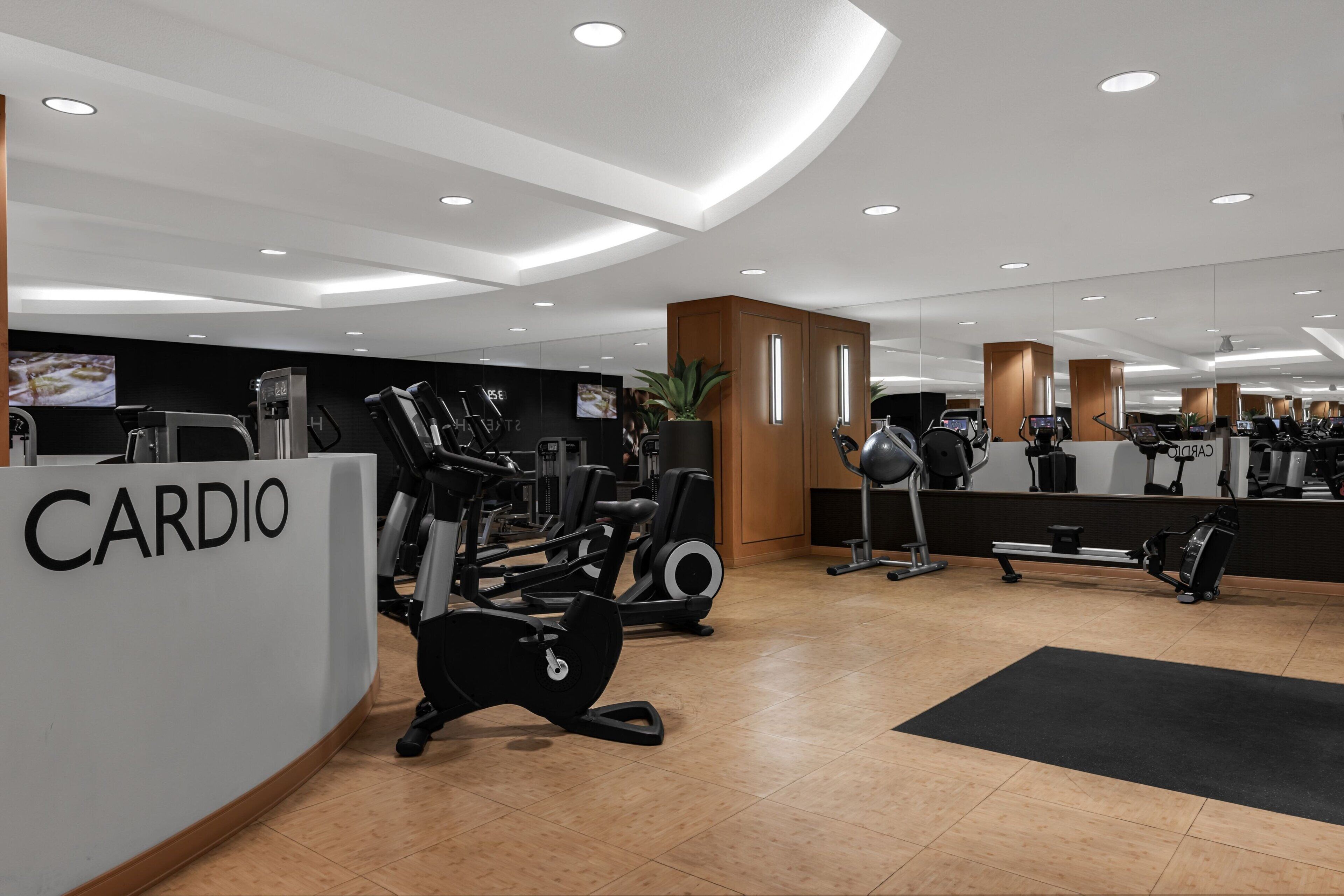 Un gimnasio moderno con equipo de cardio, un mostrador de recepción que dice 'CARDIO' y un espejo grande que refleja el espacio.