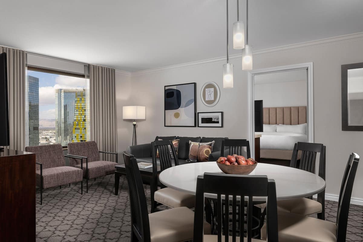 Ein Hotelzimmer mit einem runden Esstisch, einer Couch und einem Bett. Zwei Stühle stehen am Tisch, eine Lampe ist vorhanden und es gibt ein Fenster mit Vorhängen.