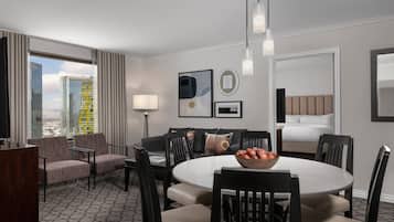 A hotel room with a round dining table, a sofa, and a bed. There are two chairs around the table, a lamp, and a window with curtains.