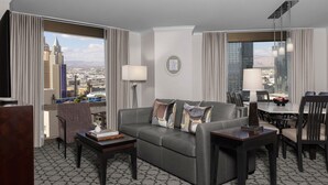 A modern hotel room with a gray patterned carpet, a gray sofa with decorative pillows, a wooden coffee table, a side table with a lamp, a dining area with a table and chairs, and a large window offering a city view. - Marriott's Grand Chateau (Las Vegas)