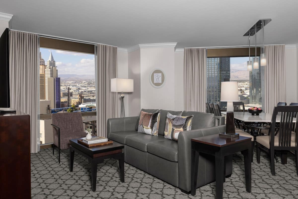 Ein modernes Hotelzimmer mit grauem Strukturteppich, einer grauen Couch mit Zierkissen, einem Holztisch, einem Beistelltisch mit Lampe, einem Essbereich mit Tisch und Stühlen sowie einem großen Fenster mit Blick auf die Stadt.