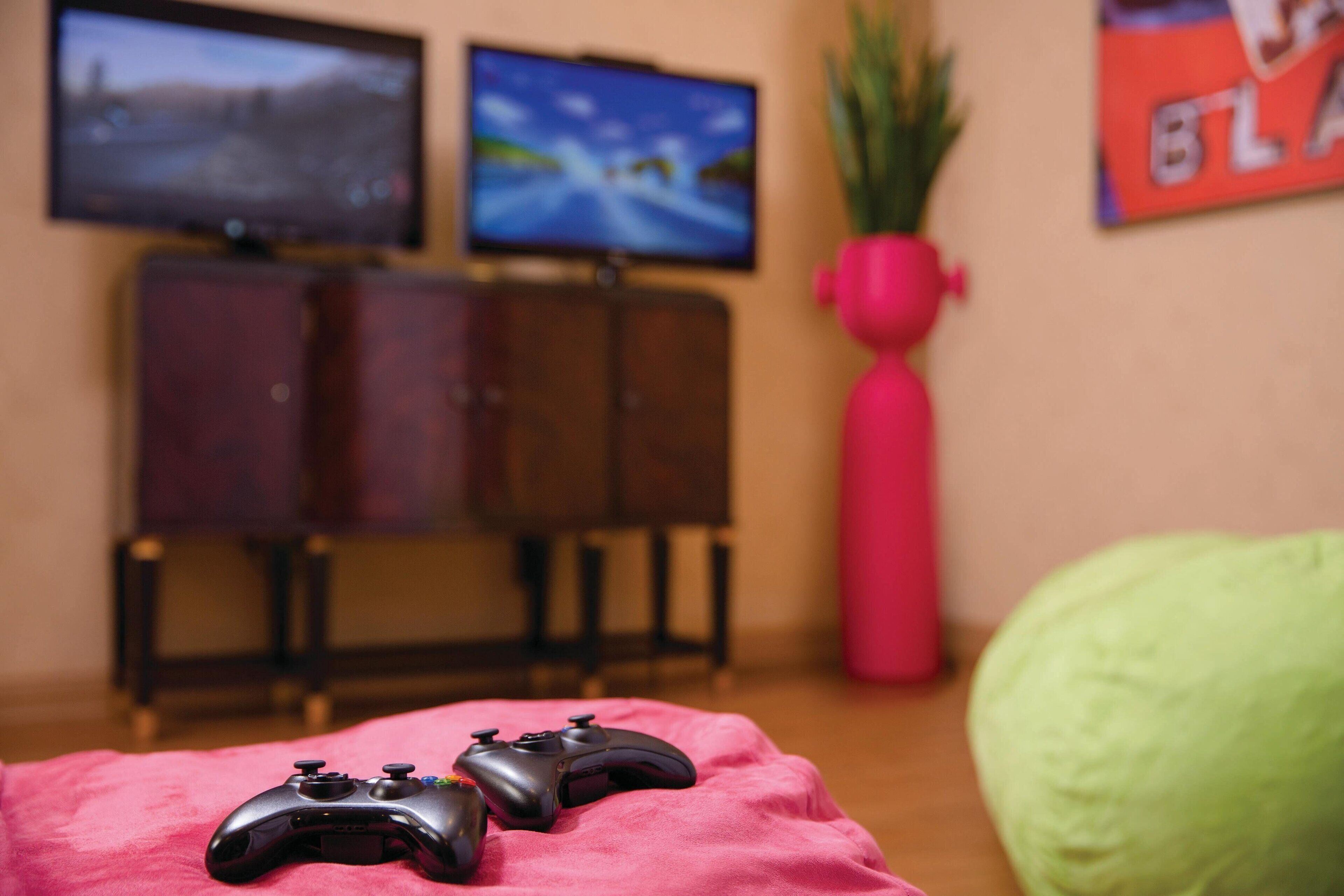 Um espaço de jogos com dois controles sobre uma manta rosa, uma TV exibindo um jogo, um armário e um vaso de planta.