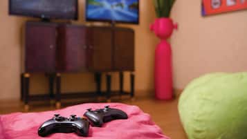 Um espaço de jogos com dois controles sobre uma manta rosa, uma TV exibindo um jogo, um armário e um vaso de planta.