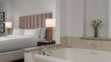 A hotel room with a bed, two bedside lamps, a nightstand, a bathroom with a bathtub, and a vase with flowers on the wall.