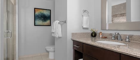 Un baño moderno con cabina de ducha de vidrio, inodoro blanco, lavabo con encimera de granito y mueble de tocador de madera oscura.