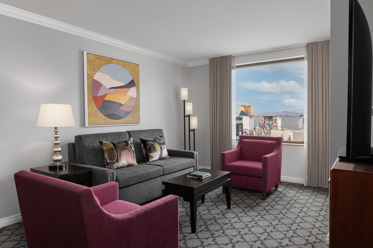 Ein Hotelzimmer mit grauem Sofa, einem roten Sessel, einem Couchtisch, einer Lampe, einer Stehlampe, einem Fernseher und einem großen Fenster mit Blick auf Gebäude.