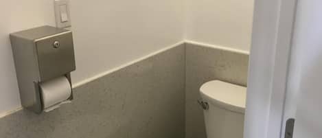 A small, compact bathroom with a toilet, a paper towel dispenser, and a wall-mounted light switch.
