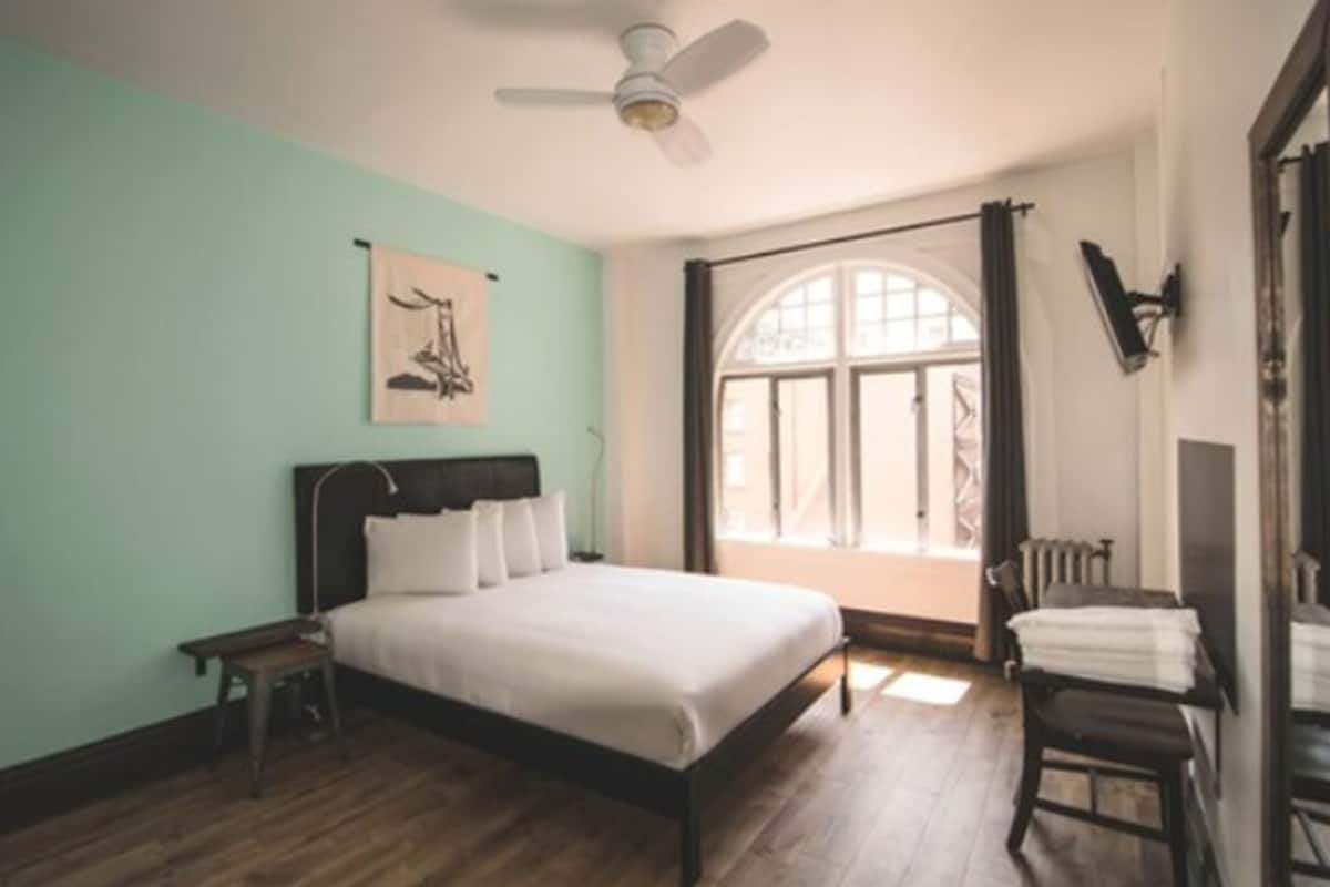 Una camera da letto con un letto, un comodino, una sedia, una finestra con tende e una ventola a soffitto.