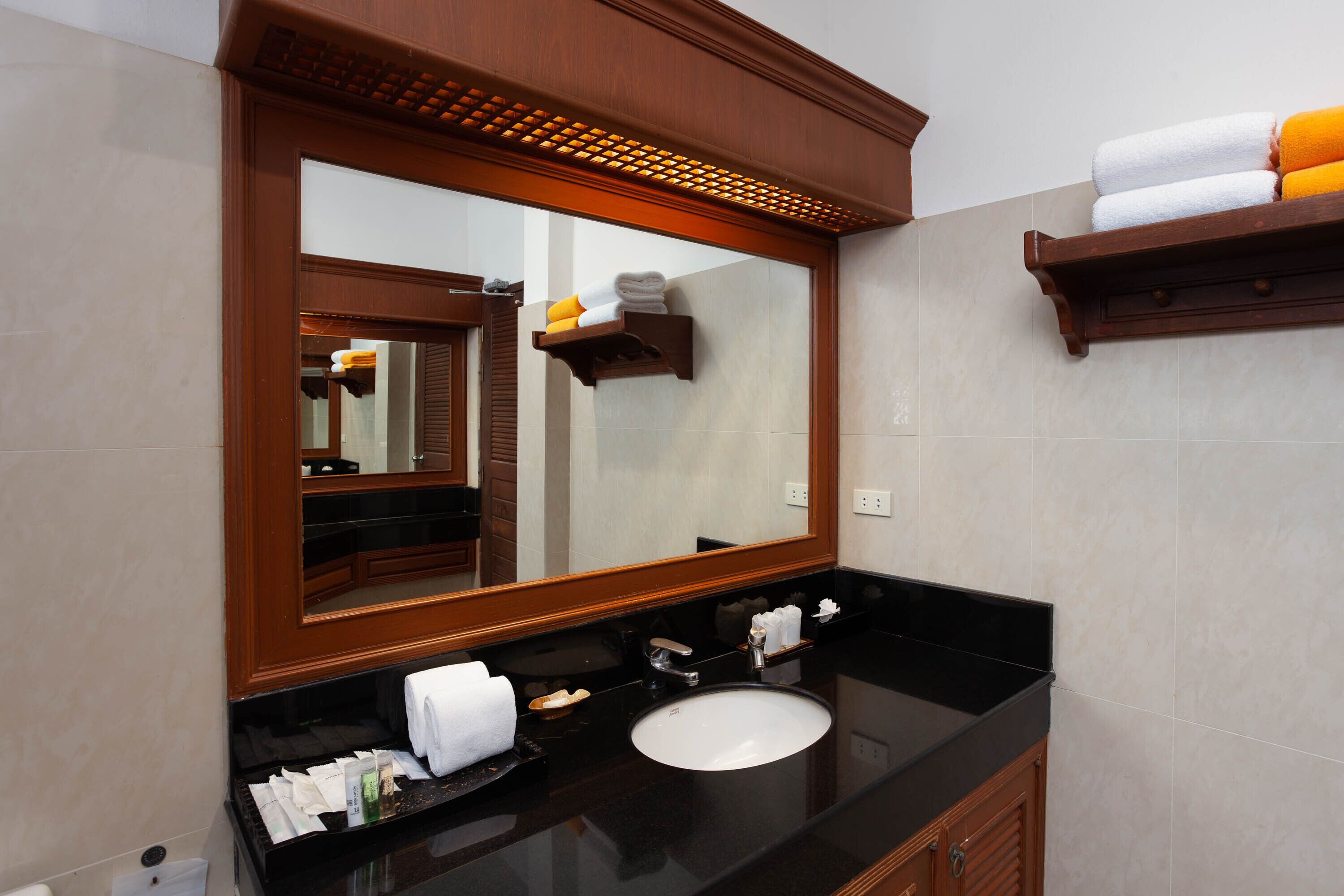 Un baño con un espejo grande, encimera negra, lavabo blanco y estantes de madera con toallas.
