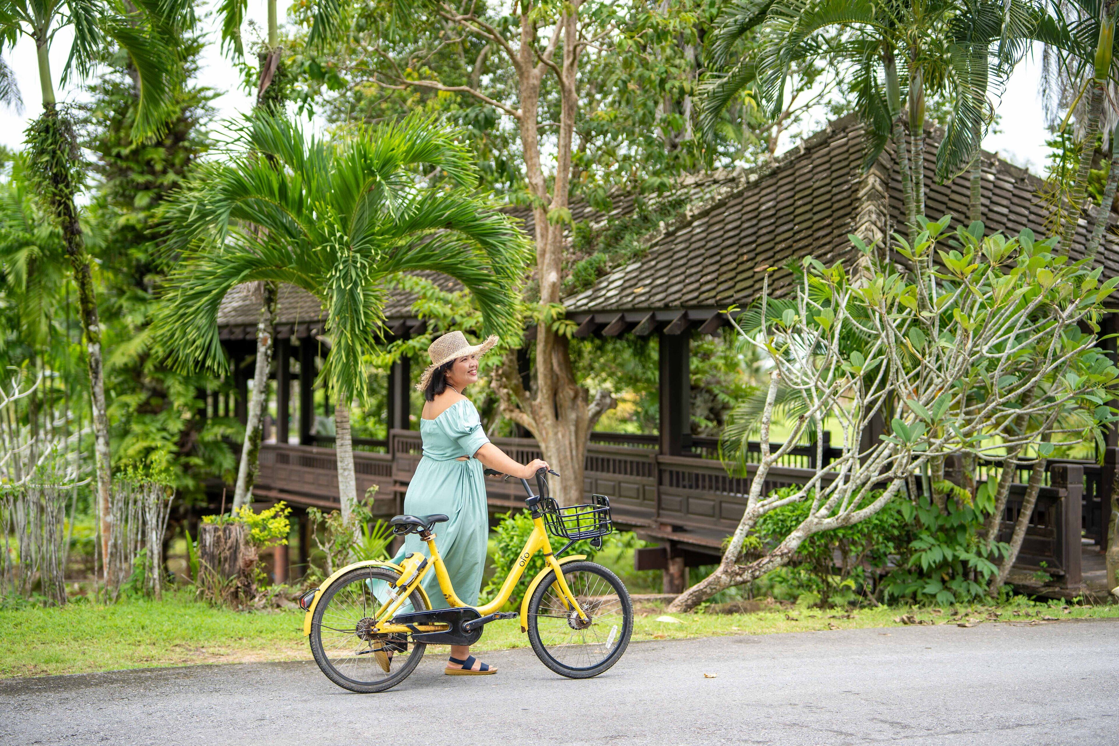 A woman in a green dress and straw hat riding a yellow bicycle with a basket, in a lush, green outdoor setting with a wooden structure in the background.