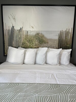 Una cama con ropa de cama y almohadas blancas, un amplio cuadro de una escena de playa y un plaid estampado al pie de la cama.