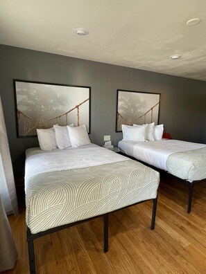 A hotel room with two beds, each with white bedding and a leaf-patterned throw. There are two framed pictures on the wall, and the room has wooden flooring. -  Valued Stay Mayflower (Madison)