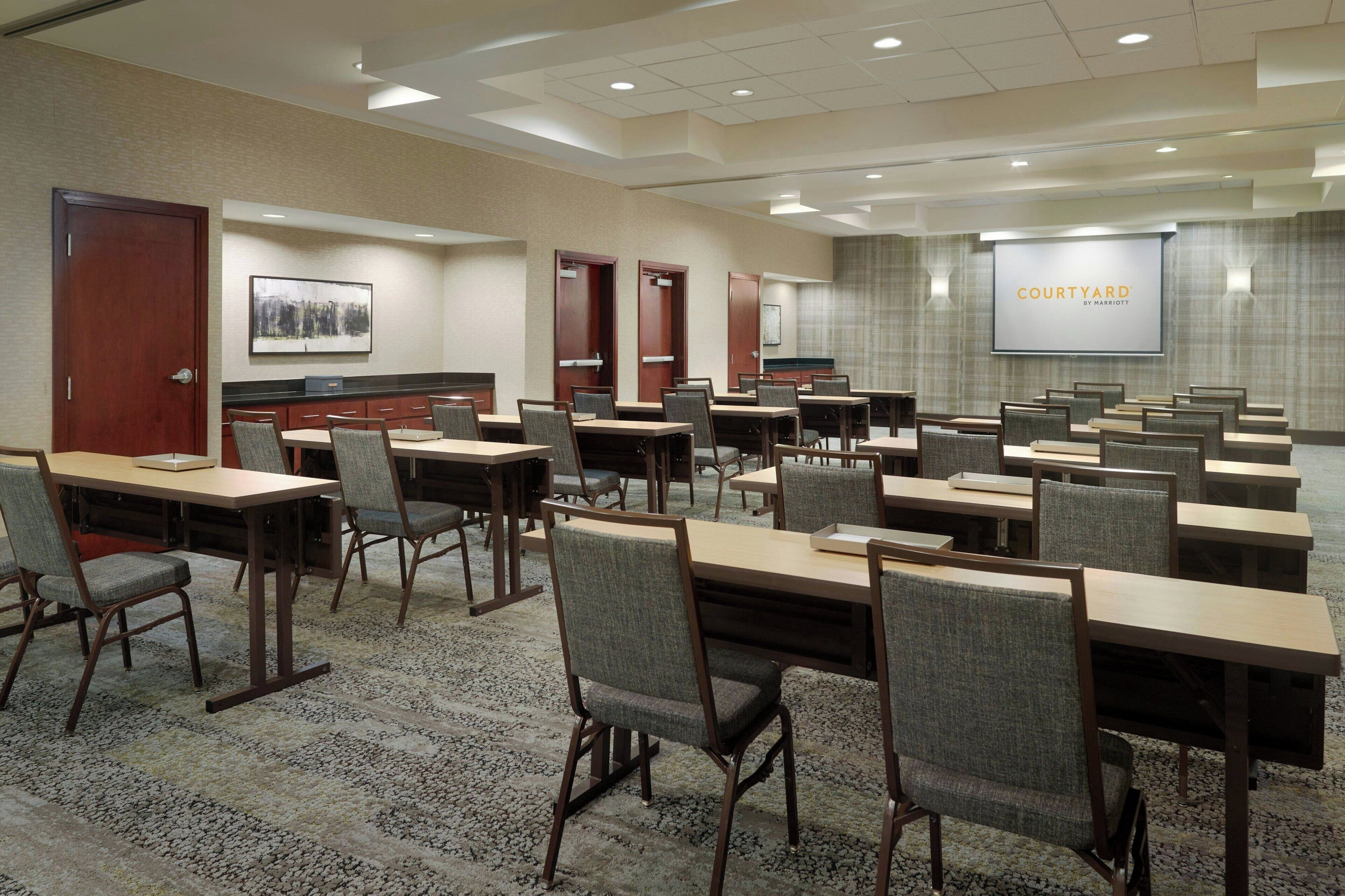 A conference room with multiple tables and chairs, a large screen displaying 'COURTYARD BY MARRIOTTE', and a carpeted floor.