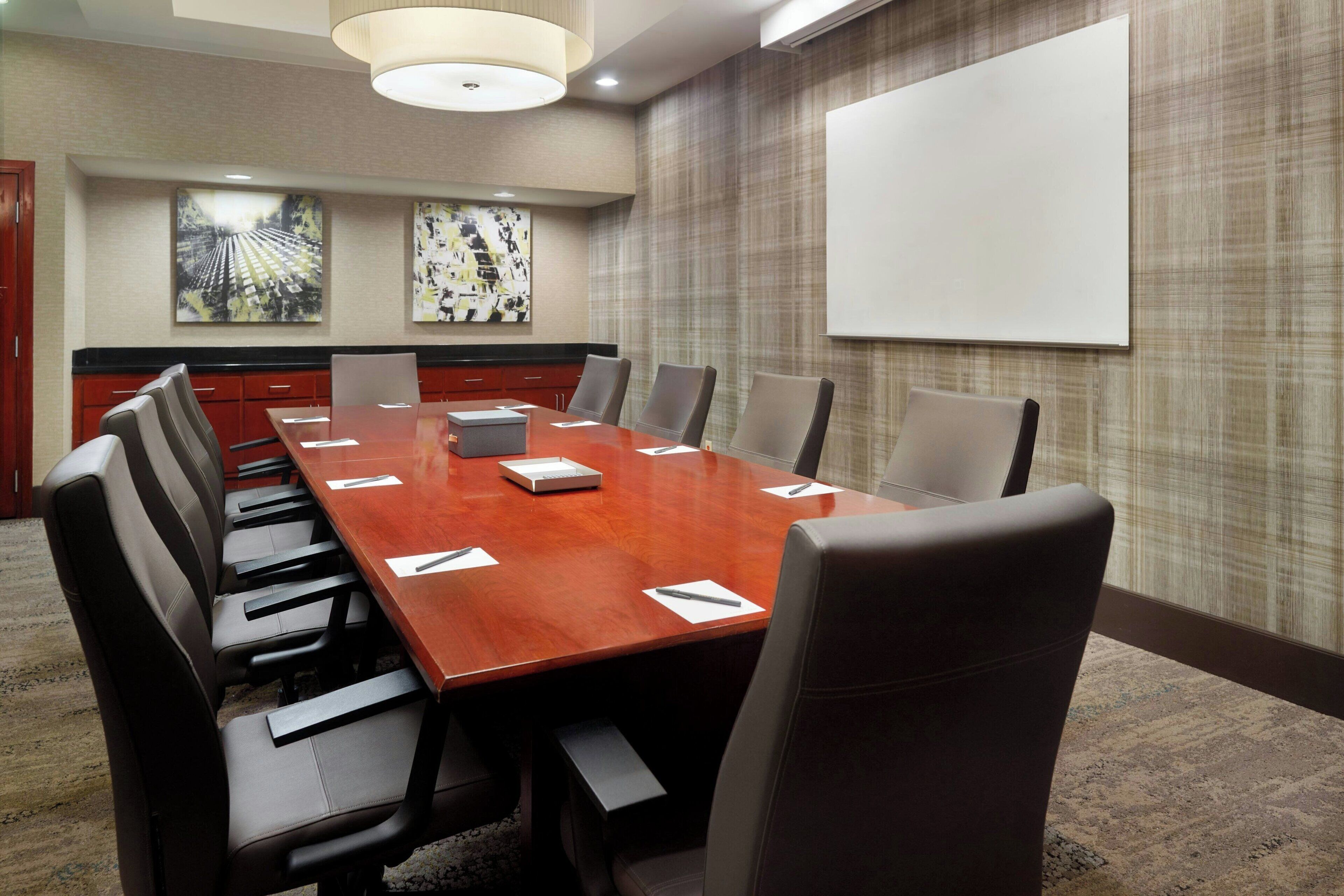 Un salón de conferencias con una mesa larga, varias sillas, una pantalla de proyector y dos cuadros abstractos en la pared.