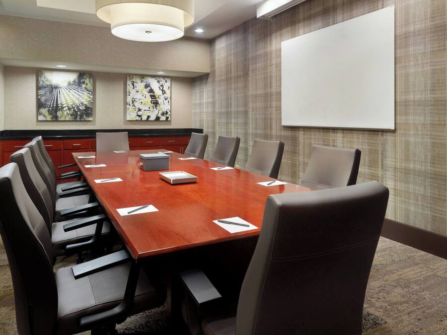 Un salón de conferencias con una mesa larga, varias sillas, una pantalla de proyector y dos cuadros abstractos en la pared.
