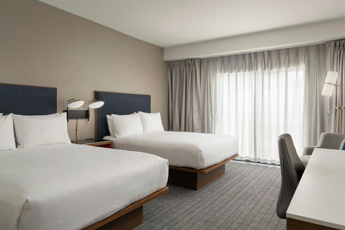 A hotel room with two beds, a desk, a chair, and a lamp.