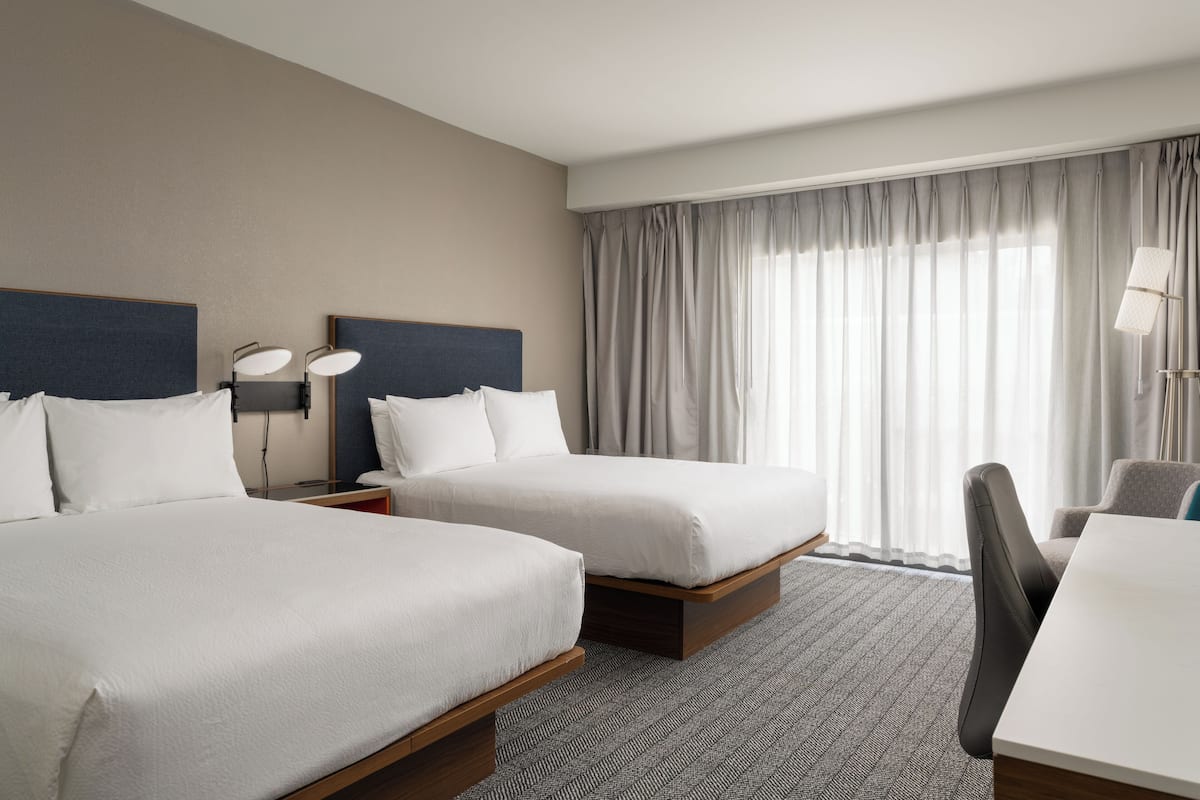A hotel room with two beds, a desk, a chair, and a large window with curtains.