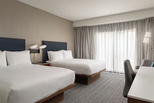 Habitación de hotel con dos camas, un escritorio, una silla y un ventanal con cortinas.