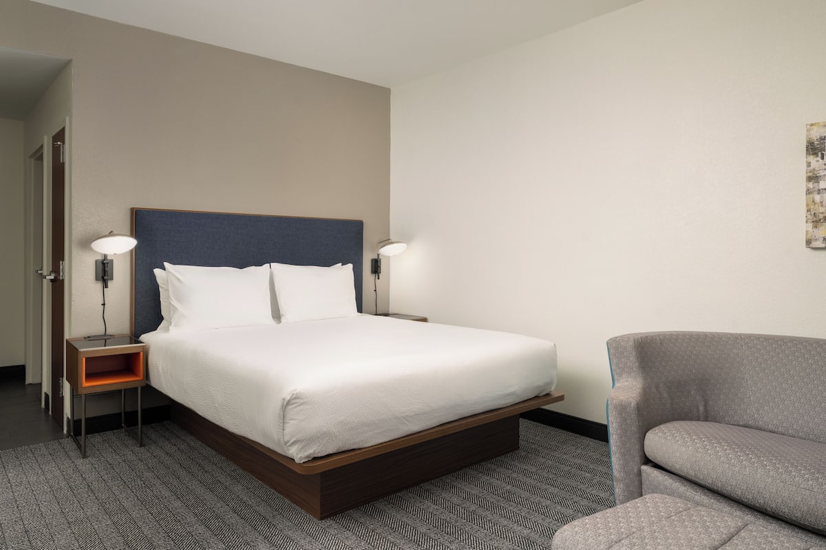 A hotel room with a large bed, two bedside tables with lamps, a gray armchair, and a patterned carpet.