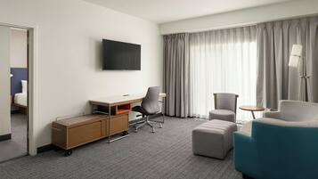 A hotel room with a desk, chair, ottoman, and a TV mounted on the wall.