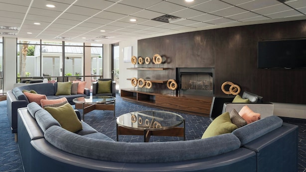 A modern lounge area with a curved sofa, a glass coffee table, and a fireplace. - Courtyard by Marriott Brunswick (Brunswick)