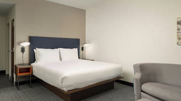 Habitación de hotel con una cama, dos mesitas de noche con lámparas, un sillón gris y una alfombra estampada.