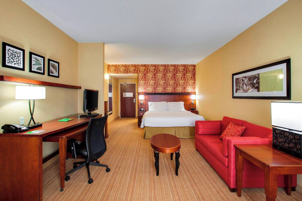 A hotel room with a bed, a desk with a computer, a red sofa, and a small table.