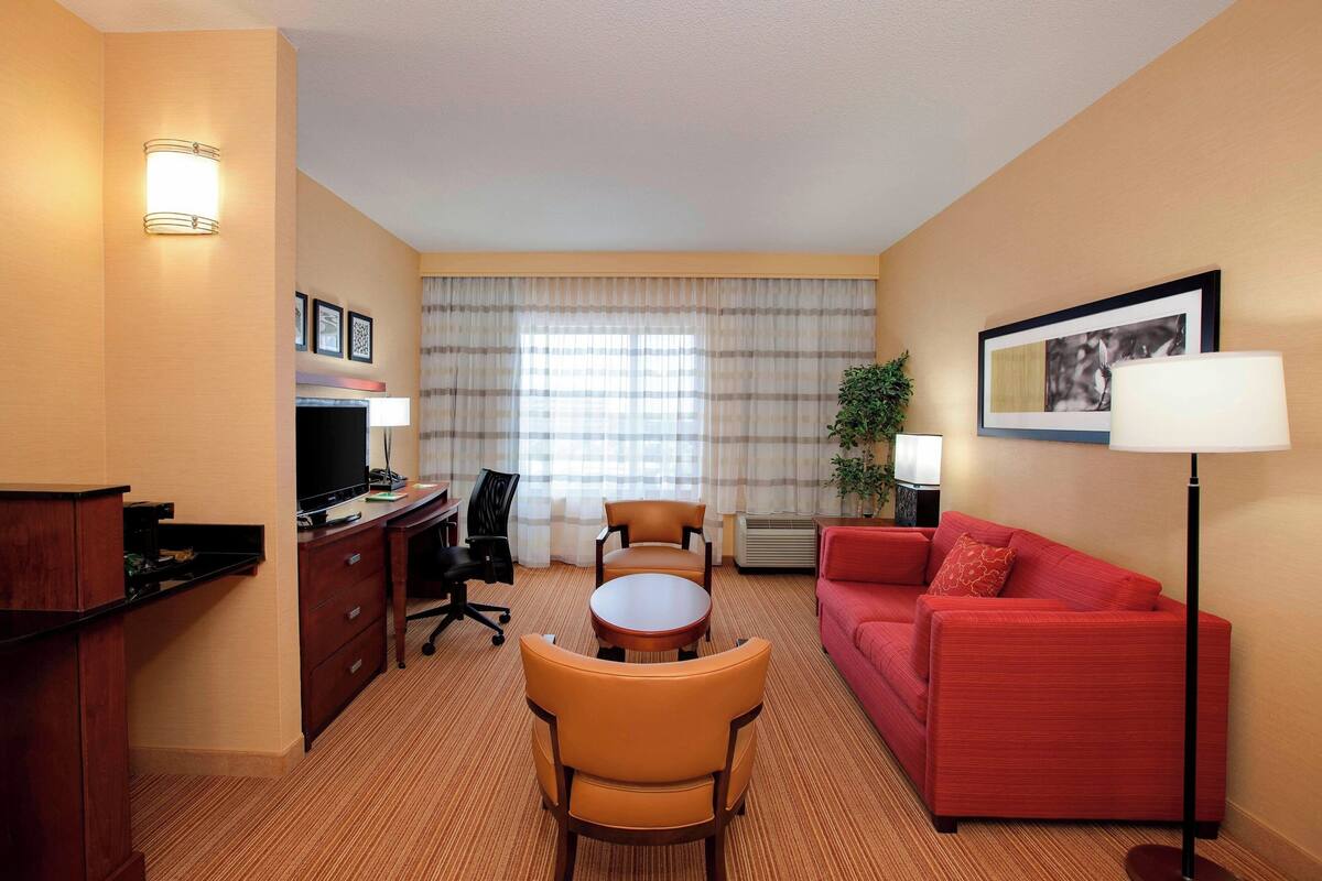A hotel room with a desk, two chairs, a red sofa, a TV, and a lamp.