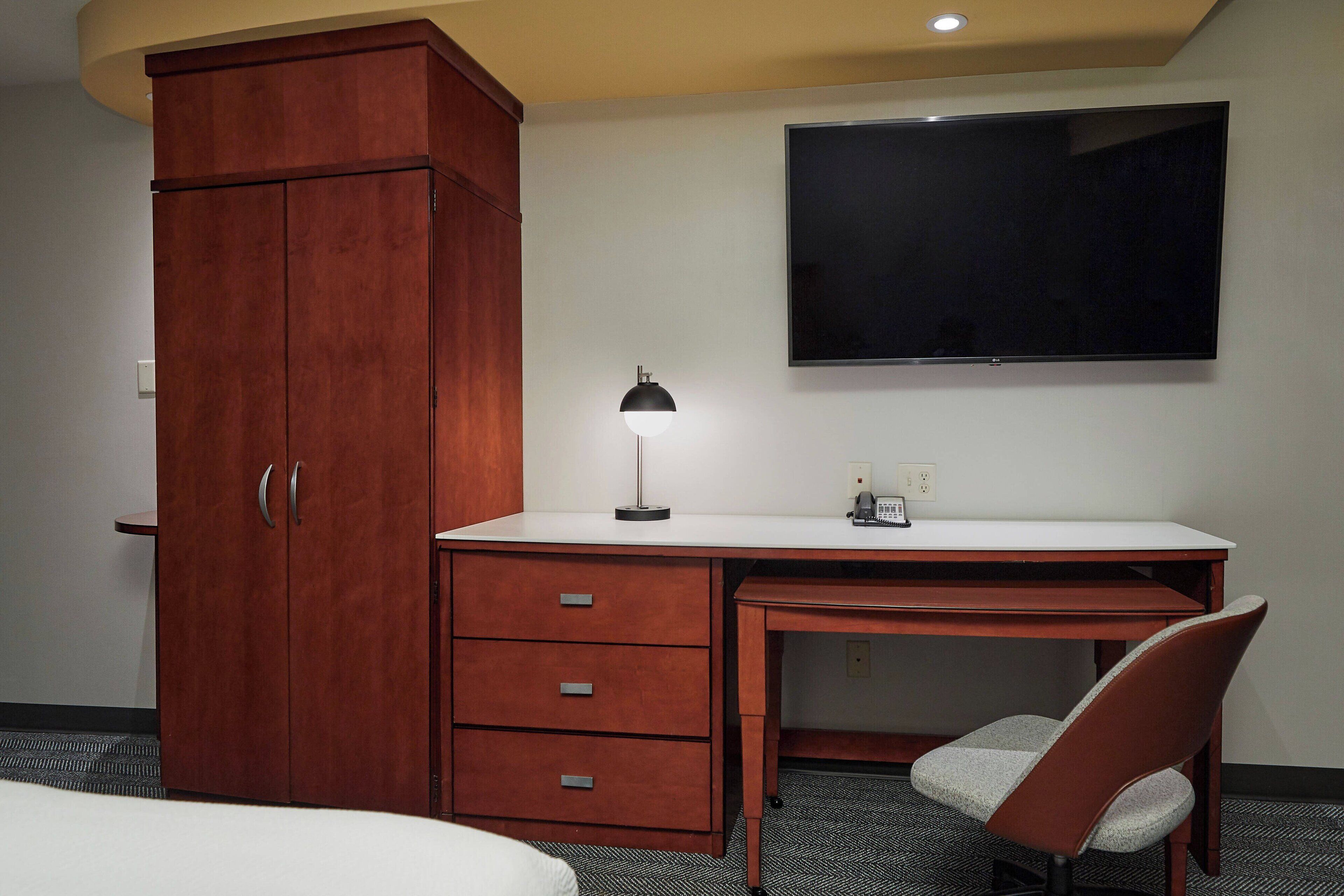 A hotel room with a wooden wardrobe, a desk with a lamp, a television, and a chair.