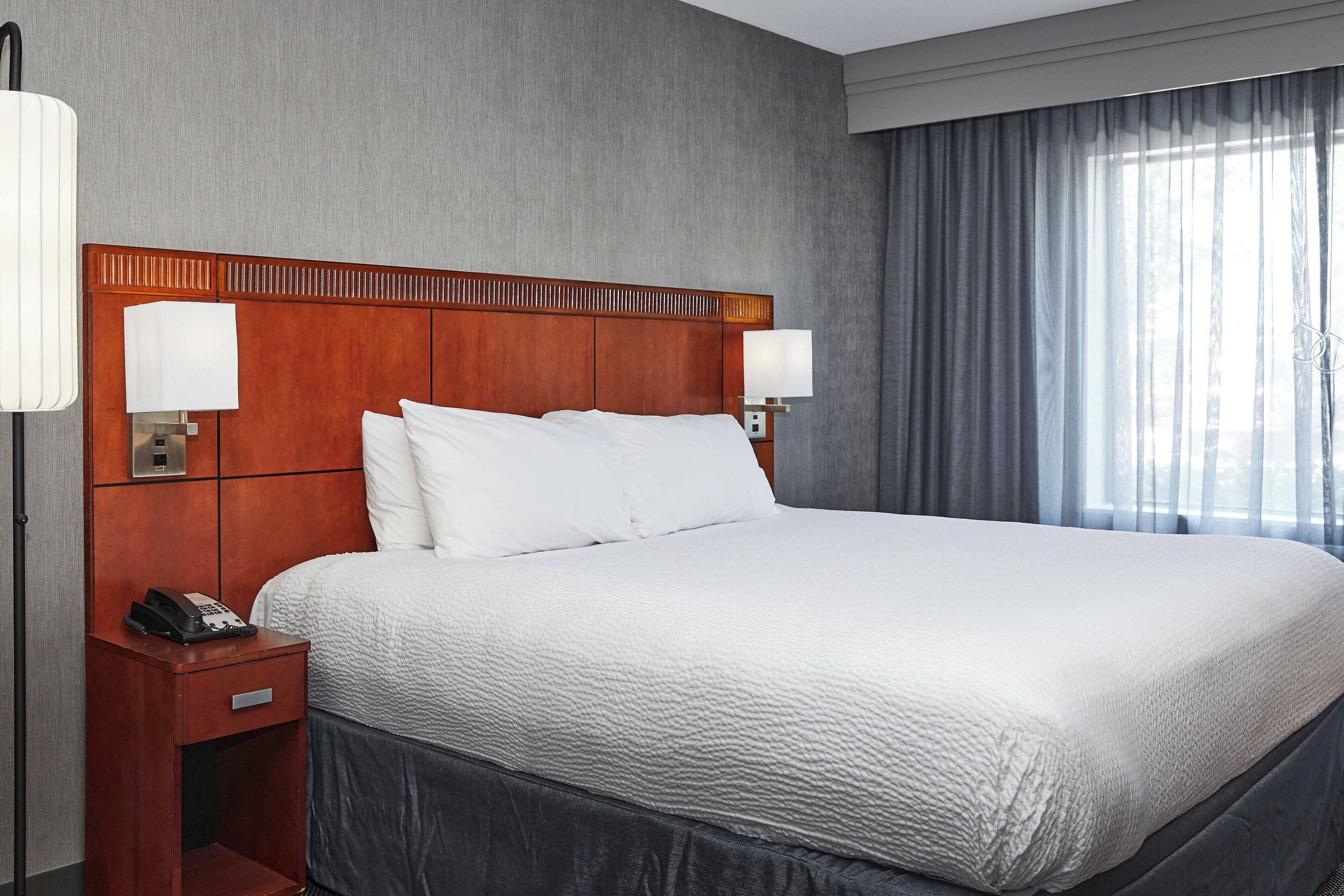 Habitación de hotel con una cama grande, cabecera de madera, dos lámparas de noche, una mesita de noche con un teléfono y una ventana con cortinas.