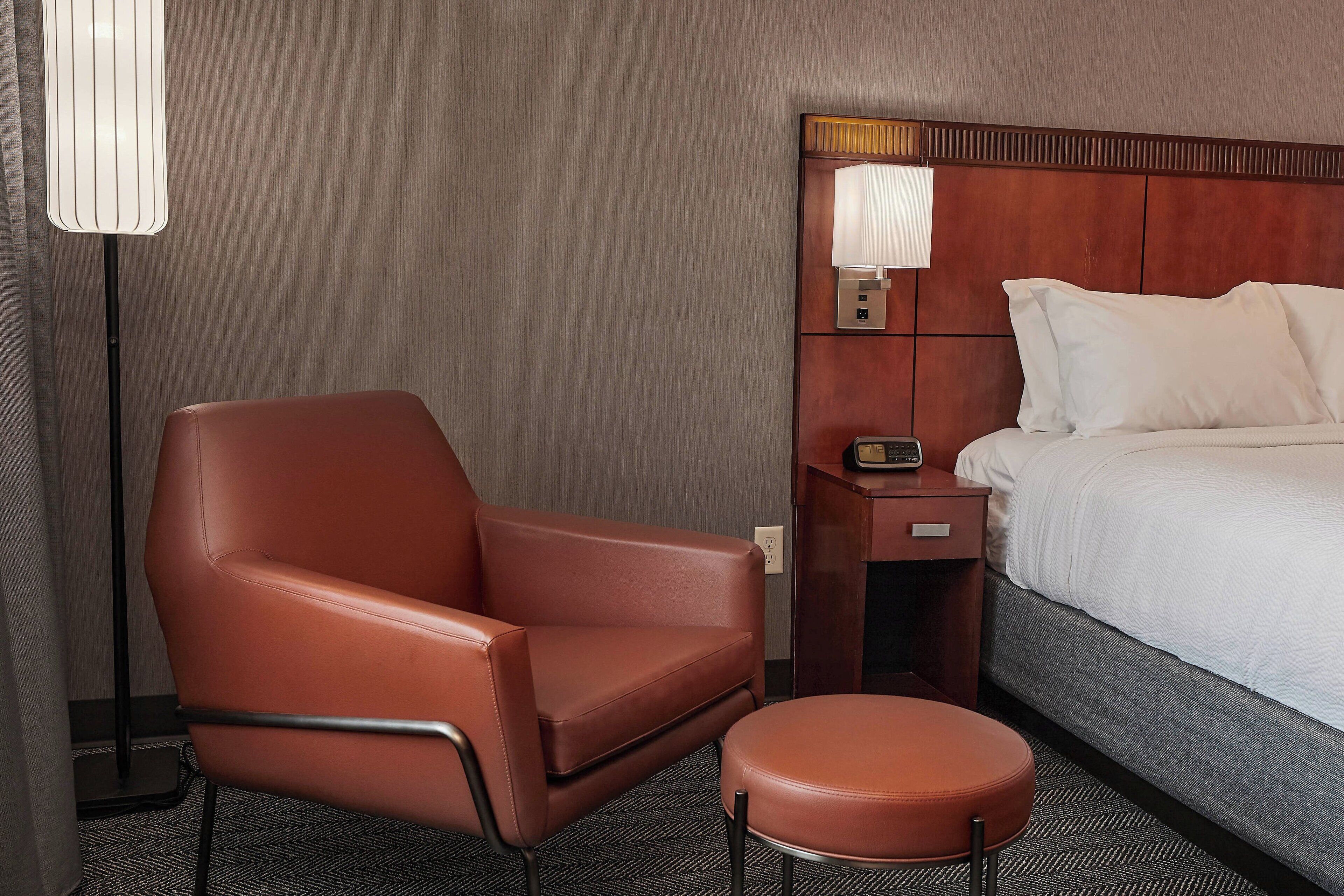 A hotel room with a bed, a brown leather chair, an ottoman, a bedside table with a lamp, and a wall-mounted control panel.
