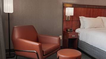 A hotel room with a bed, a brown leather chair, an ottoman, a bedside table with a lamp, and a wall-mounted control panel.