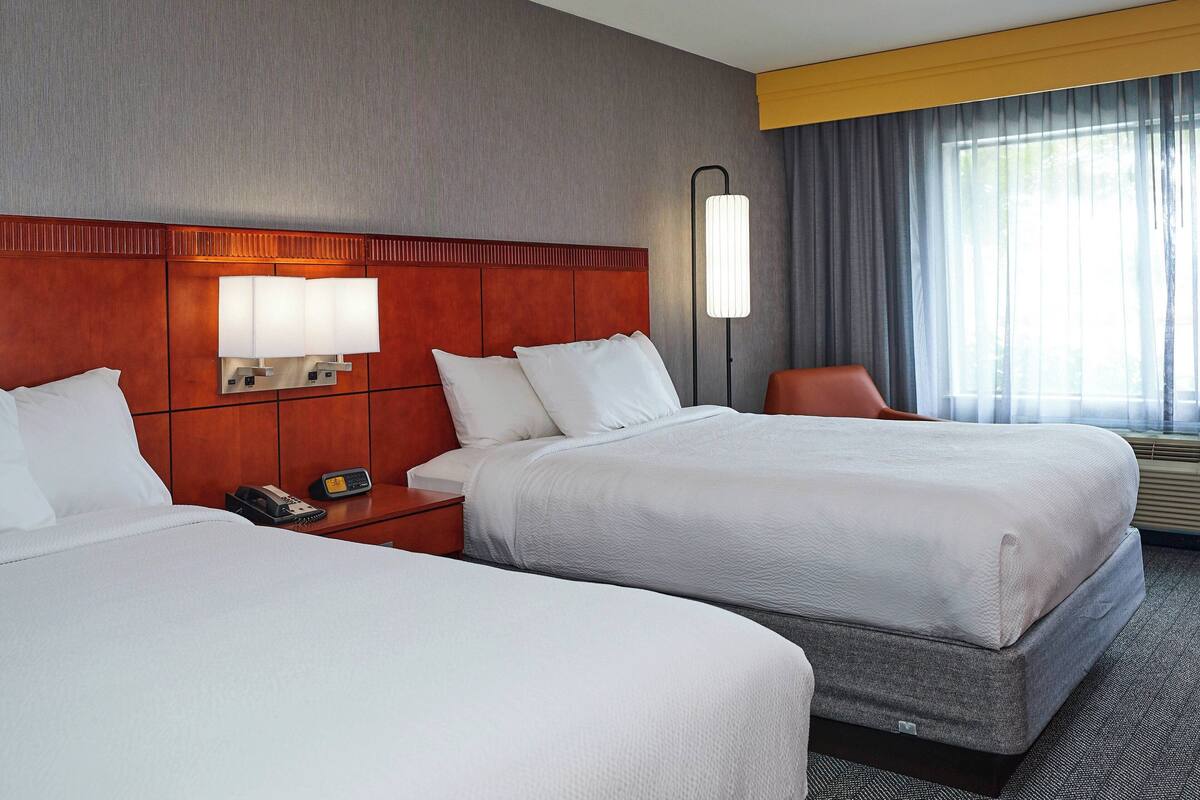 Een hotelkamer met twee bedden, een houten hoofdbord, een nachtkastje met een lamp, een stoel en een raam met gordijnen.