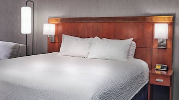 Una cama bien hecha con sábanas blancas, cabecera de madera, lámparas de noche y una mesita de noche con un reloj.