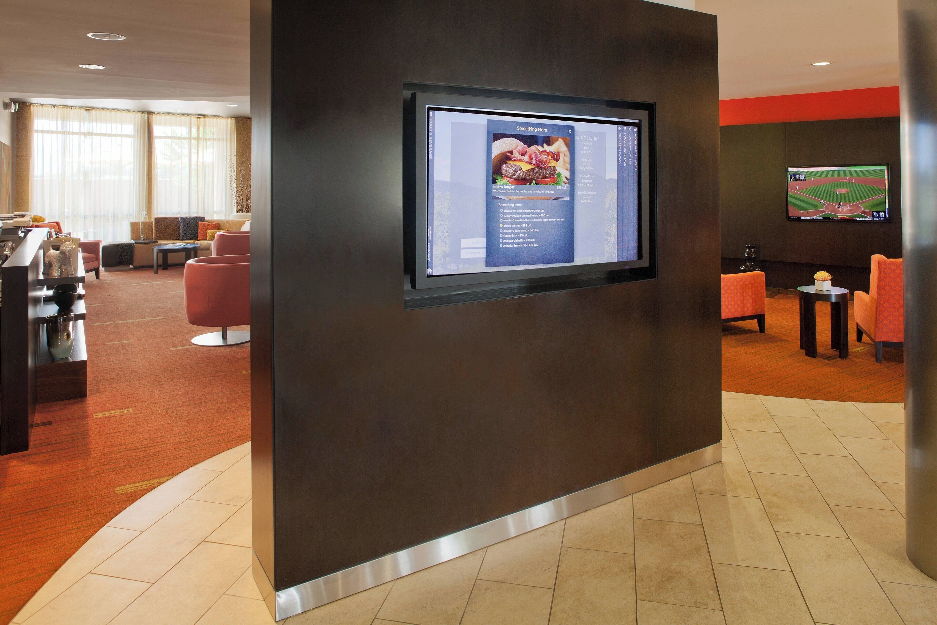 A modern hotel lobby with a large digital screen displaying a menu, comfortable seating areas, and a sports-themed wall decoration.