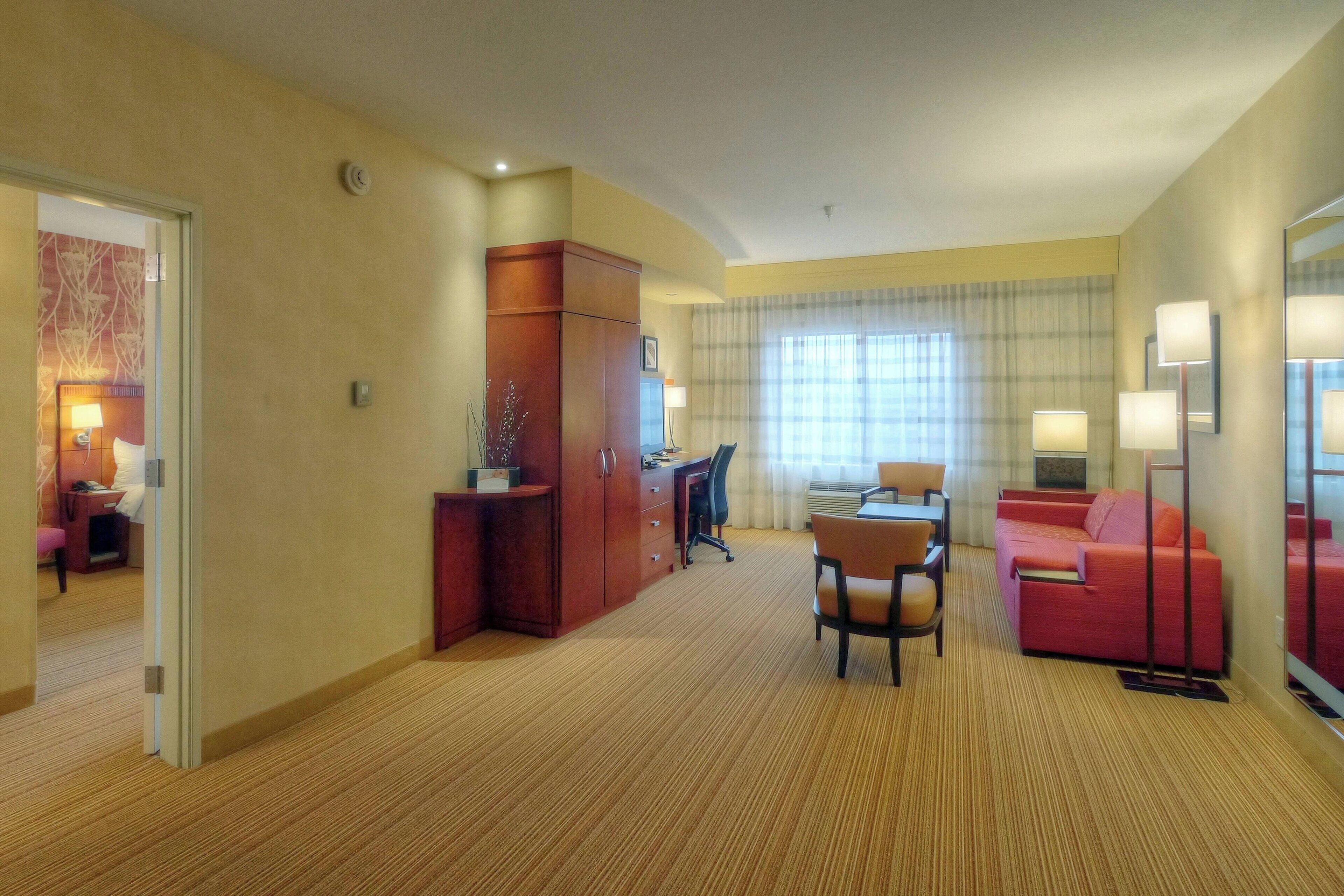 A hotel room with a carpeted floor, a desk with a chair, a sofa, and a large window with curtains.