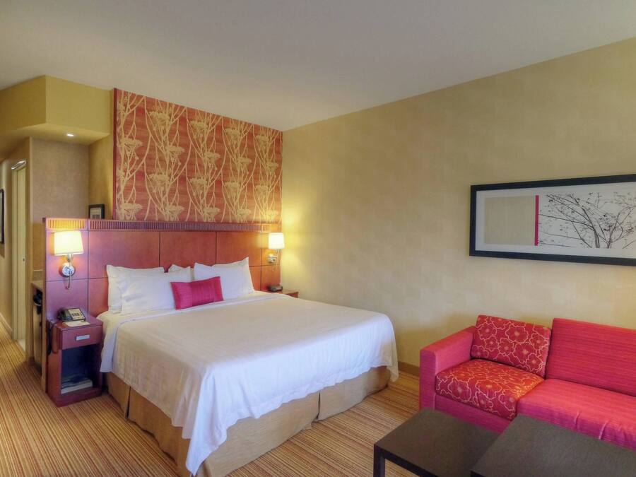 Habitación de hotel con una cama grande, un sofá rojo, una mesita de noche con lámpara y un cuadro enmarcado en la pared.