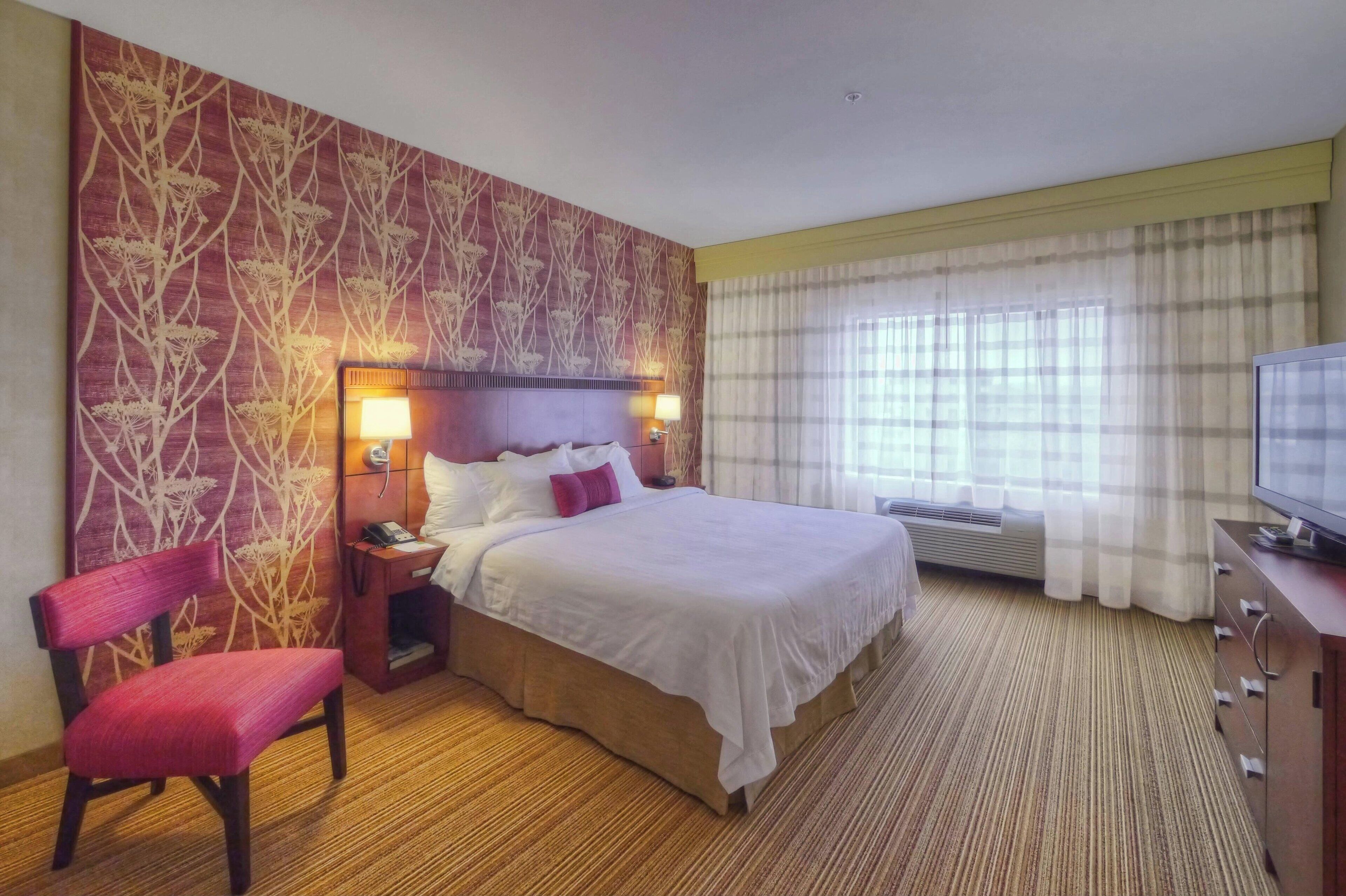Una habitación de hotel con una cama grande, una silla de madera, una mesita de noche con lámpara, un televisor y una ventana con cortinas.