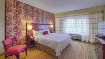 Una habitación de hotel con una cama grande, una silla de madera, una mesita de noche con lámpara, un televisor y una ventana con cortinas.