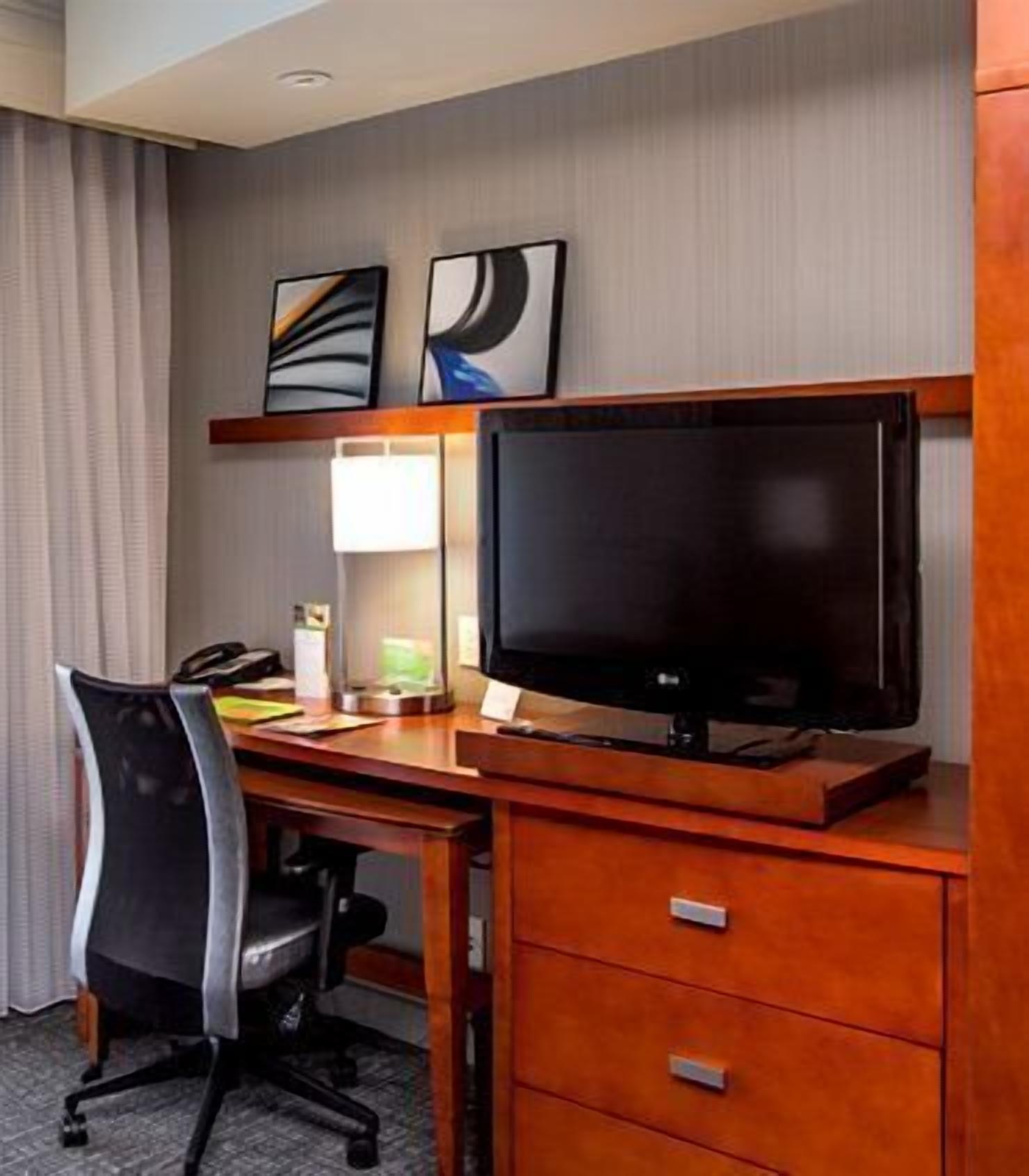 A hotel room with a wooden desk, a black office chair, a flat-screen TV, a lamp, and two framed artworks on the wall.