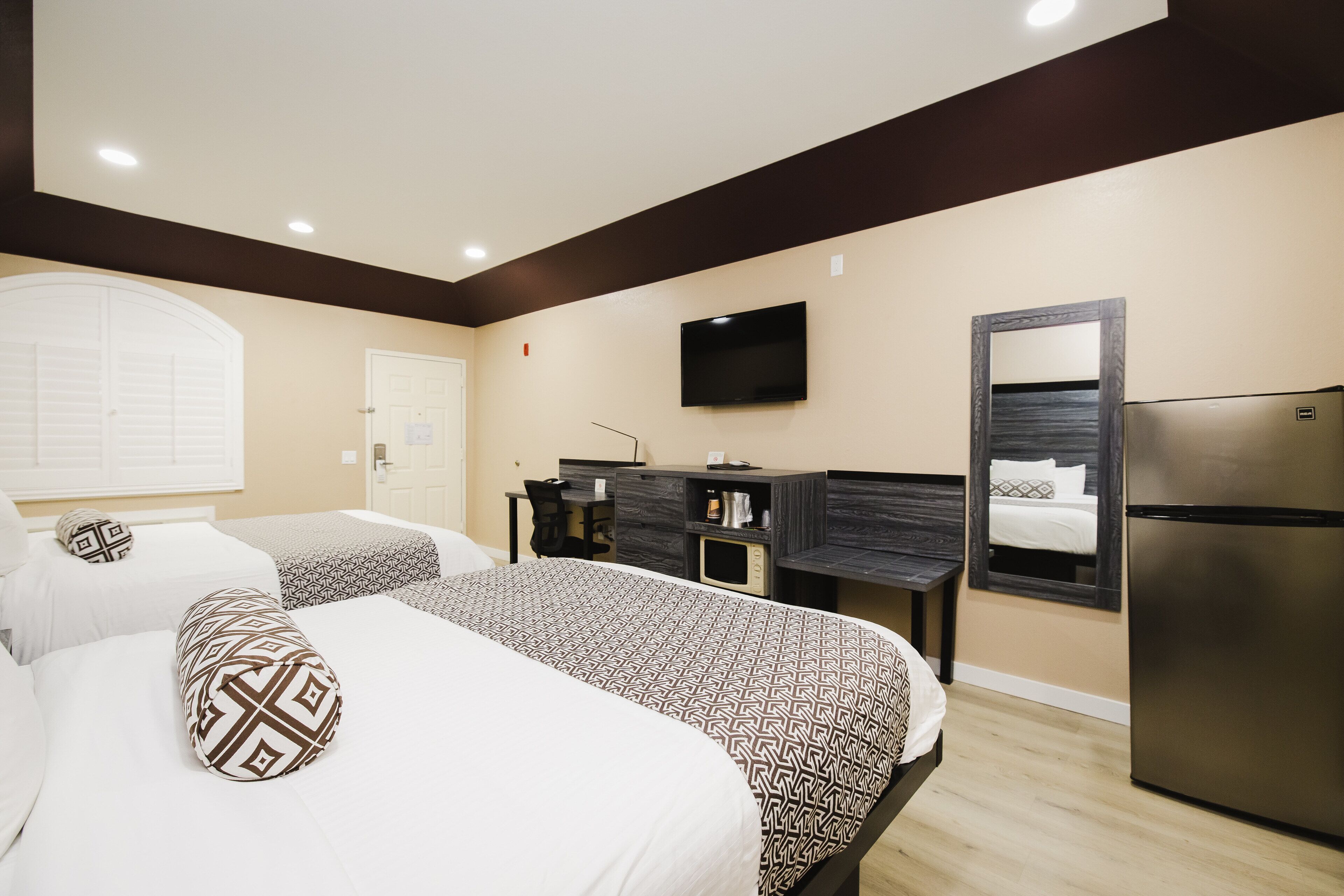 A hotel room with two beds, a TV, a mirror, a refrigerator, and a desk with a chair.
