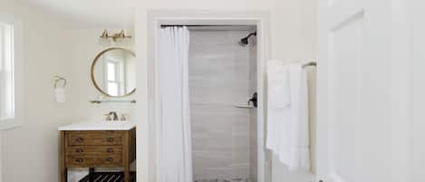 Un baño con tocador de madera, un espejo redondo, ducha con puerta de cristal y una puerta blanca.
