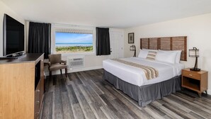 A hotel room with a bed, a desk, a chair, a TV, a window with a view of the beach, and a bedside table with a lamp. - Dune Crest Hotel (North Truro)