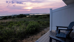 A deck with two Adirondack chairs overlooking a beach with sparse vegetation and a distant view of a body of water and a city skyline at sunset. - Dune Crest Hotel (North Truro)