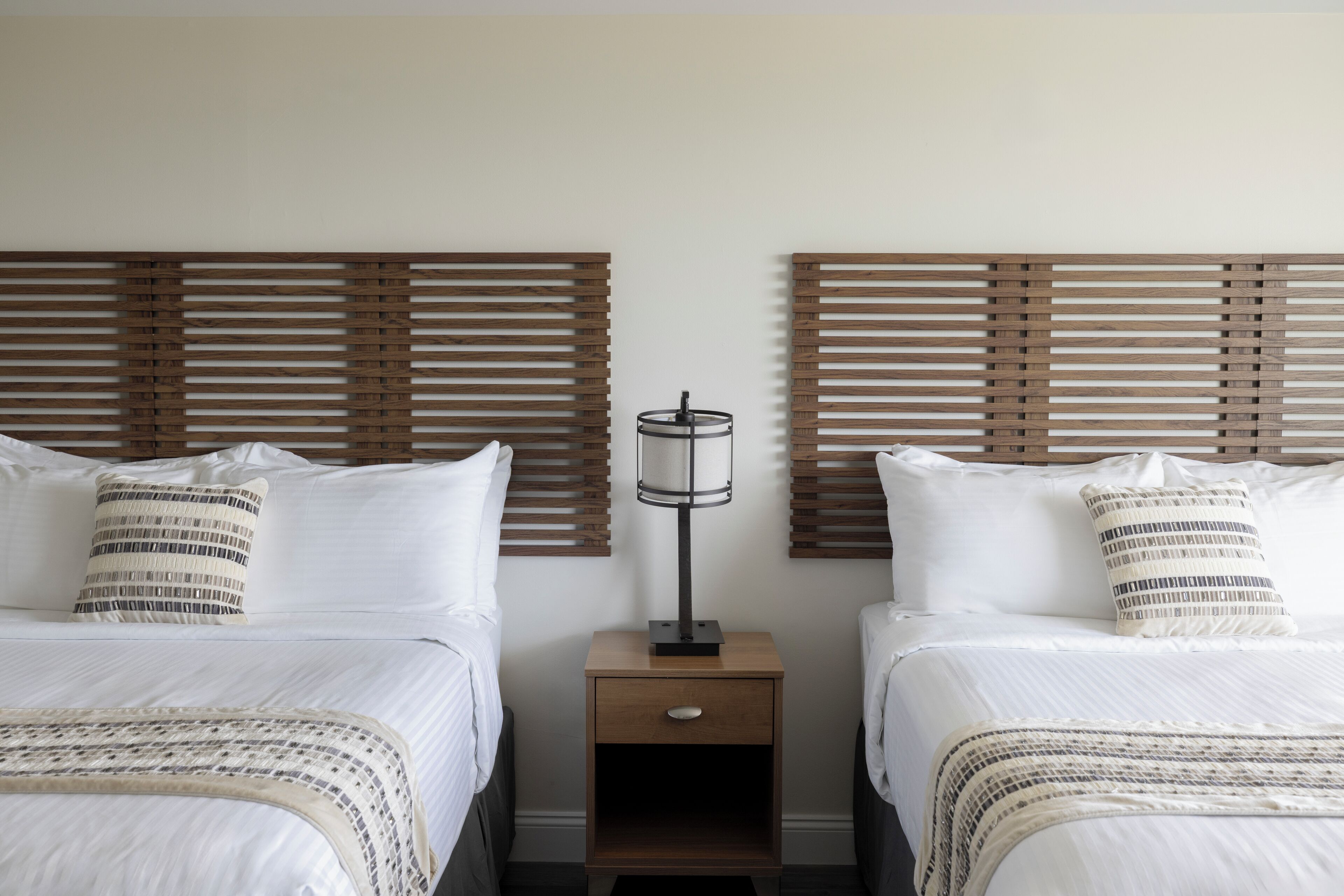 A hotel room with two beds, each with a wooden headboard, a bedside table with a lamp, and a small drawer.