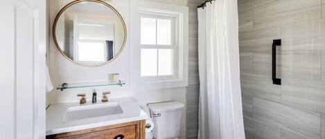 A bathroom with a white sink, wooden vanity, a round mirror, a toilet, and a shower curtain.