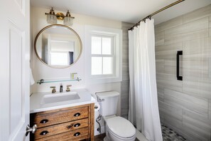 A bathroom with a white sink, wooden vanity, a round mirror, a toilet, and a shower curtain. - Dune Crest Hotel (North Truro)