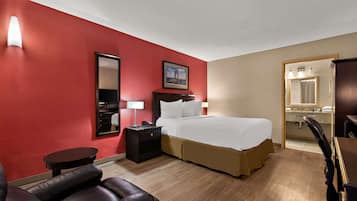 A hotel room with a red wall, a bed with white bedding, a nightstand with a lamp, a leather chair, and a desk with a chair.