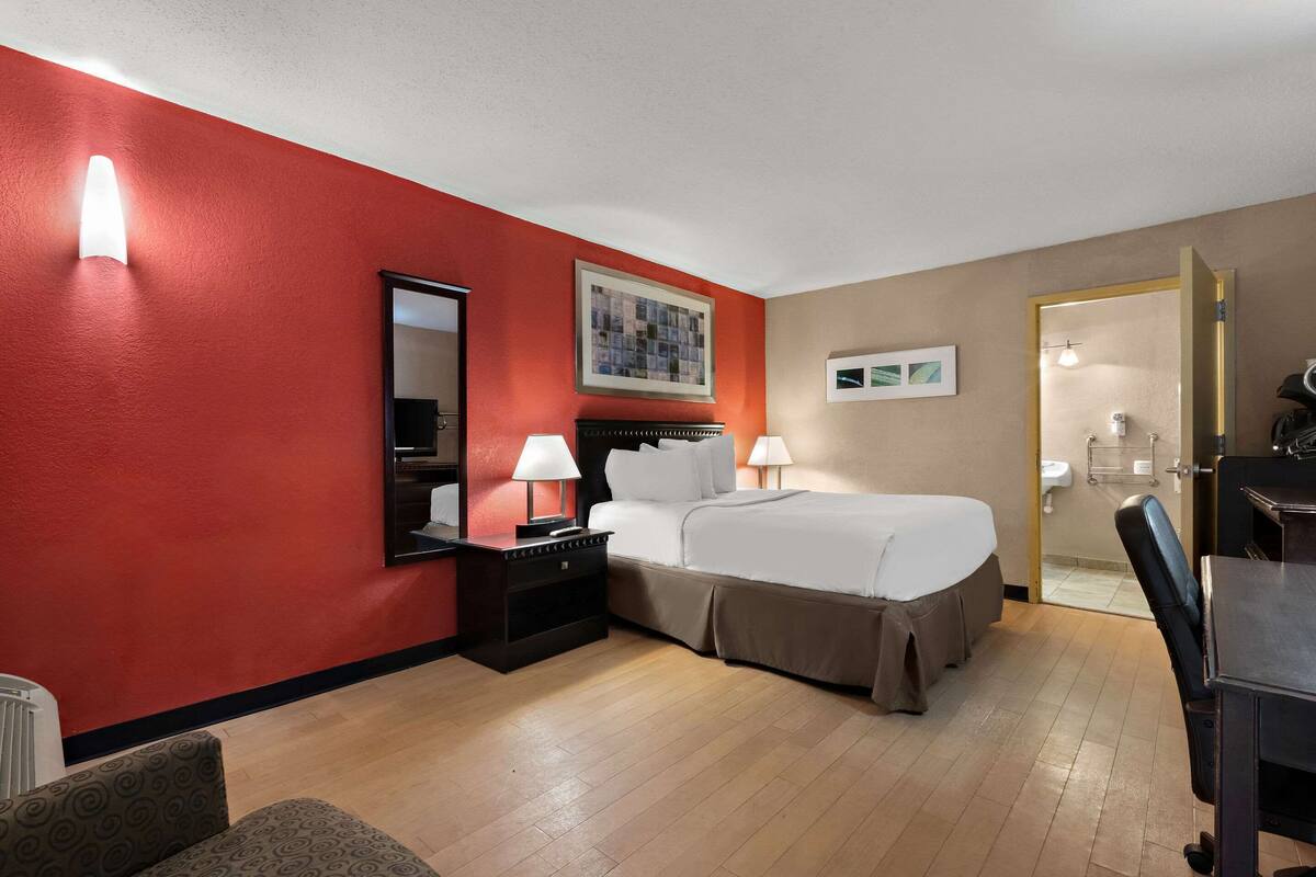 Quarto de hotel com parede vermelha de destaque, uma cama grande, duas mesas de cabeceira com luminárias, um espelho e uma porta para o banheiro.